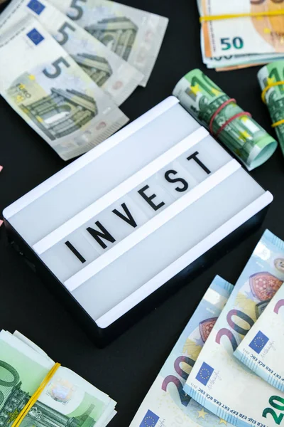 Доска с надписью INVEST черными буквами вокруг банкнот евро. Деньги, бизнес, финансы, инвестиции, сбережения — стоковое фото