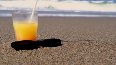 Sahilde kokteyl içeceğiz. Güneş gözlüğü, tatil, rahatlama. Yaz sahili arka planında kumsalda bir bardak smoothie suyu ve güneş gözlüğü kapat. Dalgalar tatil tatili