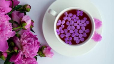 4k Rüzgârı pembe şakayık çiçekleri ve parlak masada bir fincan çay üflüyor. Bahar mevsimi sevgililer günü, kadın, anne, 8 Mart bayramı, romantik kahvaltı. Şık bir blog yazarı. Tebrik kartı