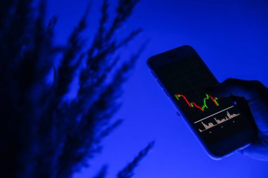 Cep telefonu tutan kadın, borsa geçmişi grafiği, kripto para birimleri ya da bitcoin konseptiyle zincire vurulmuş. Gece mavisi ışık. Ekranda genel bilgiler gösterilen gelecekteki borsa