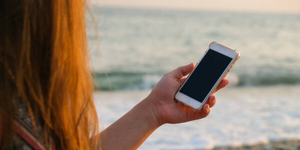 Неузнаваемая молодая женщина держит мобильный телефон на пляже против морских волн. Закат. Социальные сети, современные технологии. Летние каникулы.
