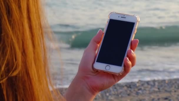 Молодая женщина смотрит на мобильный телефон на пляже у моря на закате. Зависимость от мобильных смартфонов. Всегда подключенные поколения общаться через Интернет. Чёрный экран — стоковое видео