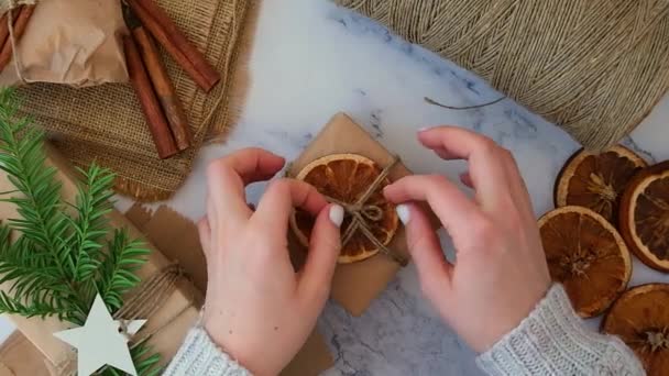 Kobieta robi pudełko z prezentów noworocznych, zawinięte w papier rzemieślniczy i ozdobione suchymi plasterkami pomarańczy. Koncepcja świąt i prezentów. Handmade Eco friendly alternatywne zielone Boże Narodzenie prezentuje zero odpadów — Wideo stockowe