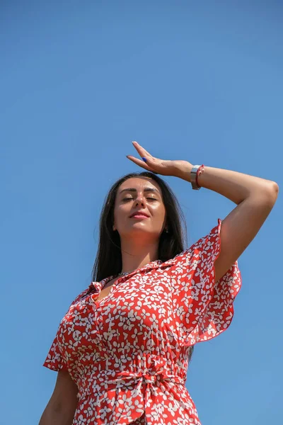 Расслабляющая молодая женщина тысячелетия в красном платье на фоне голубого неба. Улыбающаяся девушка летом веселится. Наслаждаясь солнцем — стоковое фото
