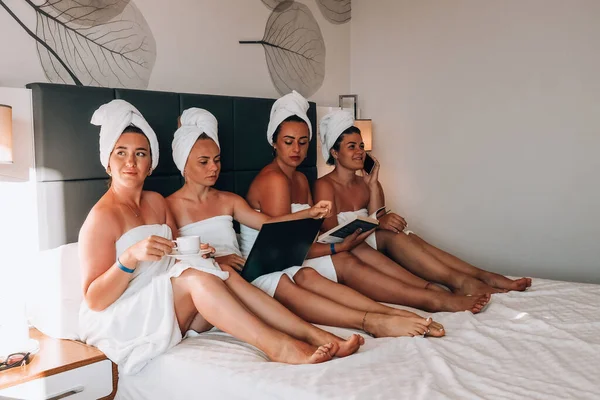 Vier meisjes vrienden in witte handdoeken zitten op het bed plezier te maken in het hotel. Vakantie Bloggers samen. Chat lief, vertel verhalen en lach. Vrienden ontmoetten elkaar en brachten hun vrije tijd door in de hotelkamer. — Stockfoto