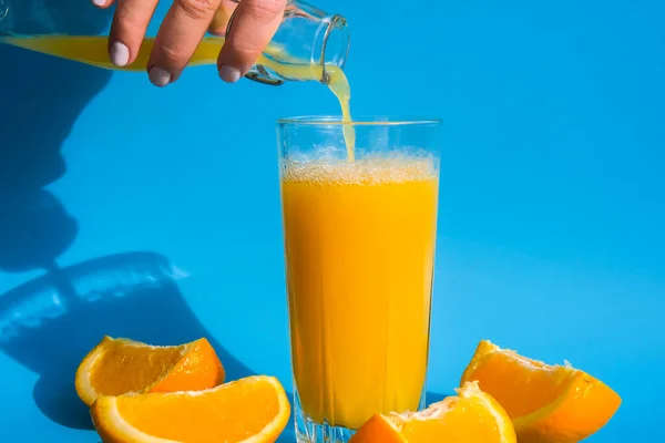 Verser le jet de jus d'orange de la cruche dans un verre de jus d'orange pressant avec des fruits tranchés sur fond bleu. Jus de fruits, boisson fraîche et fraîche. Rafraîchissements en été. Fruits coupés en deux — Photo
