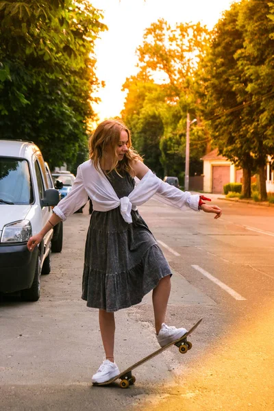 Millennial Woman in abito cavalcando uno skateboard in strada. Ragazza skater su un longboard. Fresco skateboarder femminile al tramonto. Pattinatrice spensierata 20 anni godendo della libertà stile di vita giovanile Hobby — Foto Stock