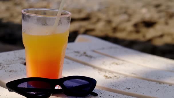 Un cóctel en la playa. Gafas de sol, vacaciones, relajado. Cerrar vaso de jugo de batido y gafas de sol en la arena en el fondo de la playa de verano. Olas vacaciones de vacaciones — Vídeos de Stock