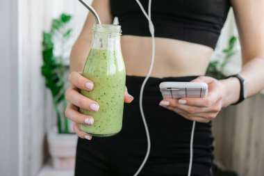 Genç bir kadın akıllı telefonu kontrol ediyor vejetaryen smoothie içiyor detoks içiyor. Evde yoga egzersizi yapan bir kadın. Çevrimiçi eğitim, temiz yemek, kilo kaybı, sağlıklı beslenme konsepti. Sağlıklı kalın. Zindelik