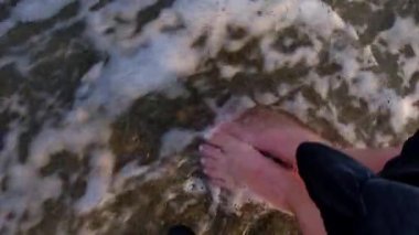 Dişi bacakları ve ayakları kumlu sahilde deniz suyu dalgaları boyunca yürüyor. Su ve köpük damlaları. Denizin kumlu sahillerinde çıplak ayakla yürüyen bir kadın..