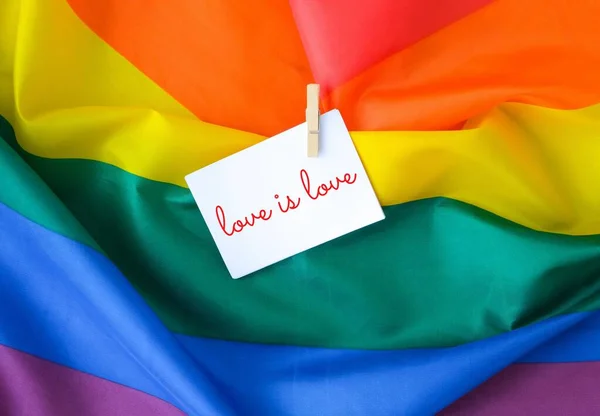 Rainbow flag with text LOVE IS LOVE message on paper note. Rainbow lgbtq flag made from silk material. Symbol of LGBTQ pride month. Equal rights. — Zdjęcie stockowe