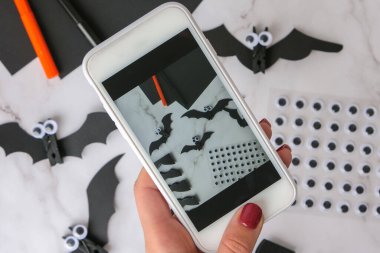 Cep telefonundan el yapımı kağıt yarasaların fotoğrafını çekiyor. Cadılar Bayramı süslemeleri. Mutlu Cadılar Bayramı partisi konsepti. Çocuk sanat projesi. Diy konsepti. Adım adım Zanaat
