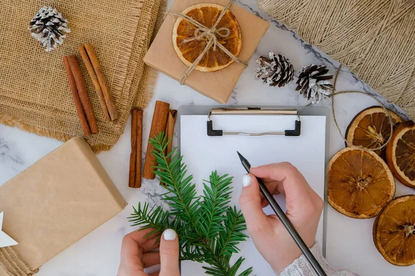 Noel tatiline hazırlanan bir kadın. Hediye listesi yazıyorum. Yeni yıl hediyeleriyle dolu bir kutu, el işi kağıdına sarılmış ve köknar dalı, kuru portakal dilimleri, tarçın çubukları ile süslenmiş. El yapımı çevre dostu alternatif yeşil Noel sunar sıfır atık Sust