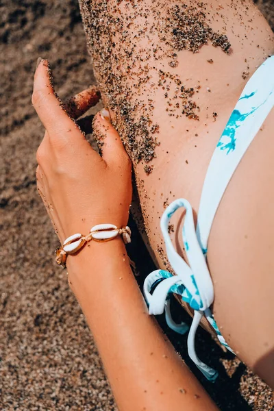 Woman\'s hands with shell jewelry in sand. Sand in hands on the beach. Travel and vacation concept. Female slim legs. Summertime relax