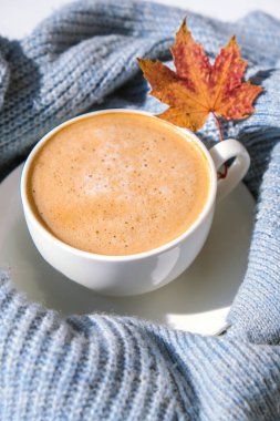 Akçaağaç sarısı yapraklı mavi örgü süveterin üzerinde beyaz bir sabah kahvesi. Konforlu ev konsepti. Estetik blog yaşam tarzı. Sonbahar sakin bir hayat. Cappuccino ya da latte sıcak içecek. Atmosfer