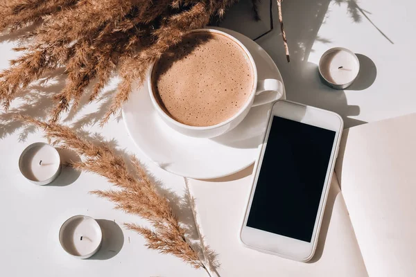 Pampas grass and white cup with coffee. Mobile phone with black screen. Checking social media Drinking Cappuccino in the breakfast morning at home. Flat lay. Wallpaper. Aesthetics Cozy home workplace