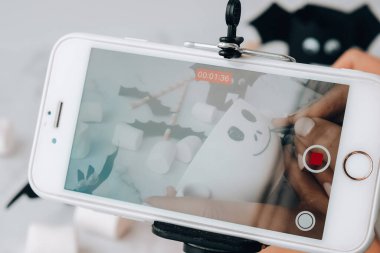 Kiev Ukrayna - Ekim 2021 Kadın elleri beyaz bardağa korkunç yüz resmi çiziyor. Çocuk blogcu cep telefonlu cadılar bayramı aktiviteleriyle video çekiyor. Tatil sanatı çocukları el yapımı dekorasyonlar yapıyor. Sanat sınıfı için ev içi fikirlere mesafeli..