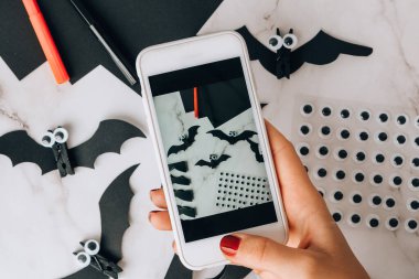Cep telefonundan el yapımı kağıt yarasaların fotoğrafını çekiyor. Cadılar Bayramı süslemeleri. Mutlu Cadılar Bayramı partisi konsepti. Çocuk sanat projesi. Diy konsepti. Adım adım Zanaat