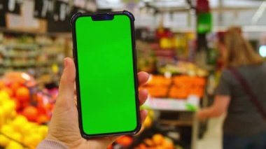 4k cep telefonu Chroma anahtarı. Alışveriş merkezinde elinde yeşil ekran olan akıllı telefonları olan kadınların yakın çekimleri. Arkaplanda süpermarketten yiyecek satın alan odaksız kadın, çevrimiçi manav merkezi