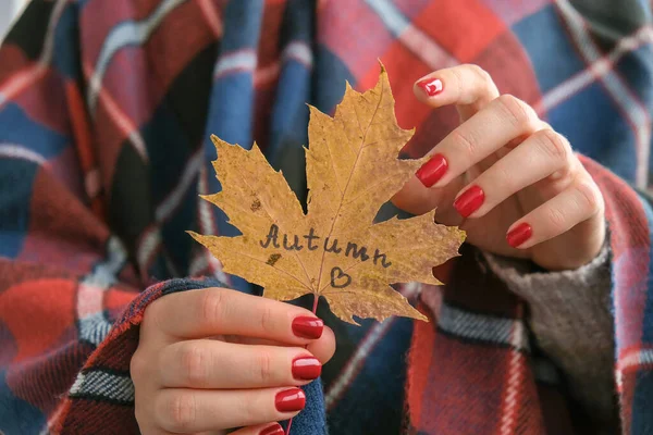 Stylish red female nails. Fall leaf with text AUTUMN in hands. Modern Beautiful manicure. Autumn winter nail design concept of beauty treatment. Gel nails. Skin care. Wellness. Trendy colors.