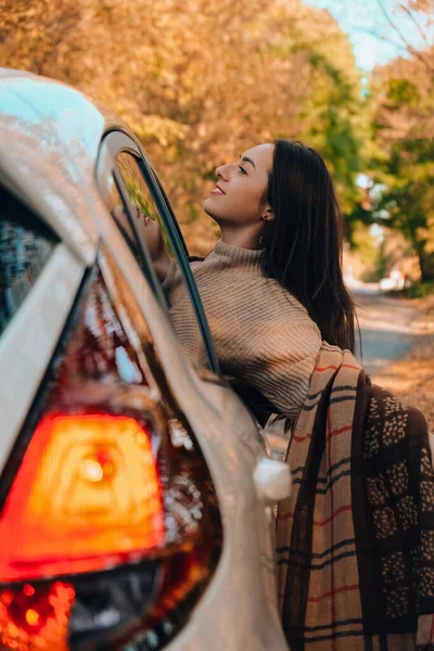 Brunette European looking cute girl sitting in a car enjoying sunny autumn weekend. Travel and road trip. Woman looking at the view from a car. Happy woman enjoying road trip. Fall Autumn forest in