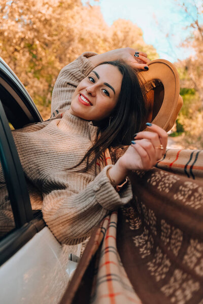 Brunette European looking cute girl sitting in a car enjoying sunny autumn weekend. Travel and road trip. Woman looking at the view from a car. Happy woman enjoying road trip. Fall Autumn forest in