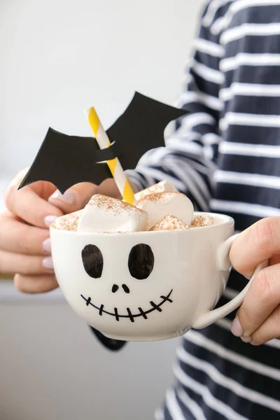 Woman holding Painted white cup scary jacks face with marshmallows Hot chocolate cacao drink DIY for kids Halloween home activities. Holiday art children craft Handmade decorations home Idea for art class.