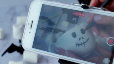 Kiev Ukraine - October 2021 Female hands paint on white cup scary face DIY for kids Blogger making video through mobile phone Halloween home activities. Holiday art children craft Handmade decorations