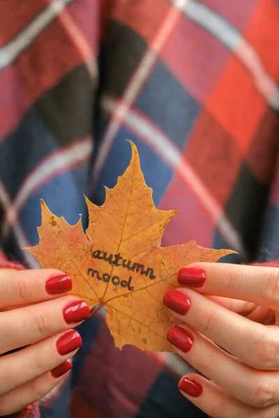 Stylish red female nails. Fall leaf with text AUTUMN MOOD in hands. Modern Beautiful manicure. Autumn winter nail design concept of beauty treatment. Gel nails. Skin care. Wellness. Trendy colors.