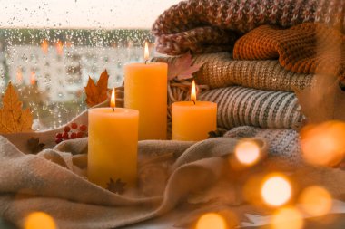 Merhaba güz, kış tatilini pencerenin kenarındaki evde kutluyoruz Şükran Günü atmosferinde Şükran Günü yaprakları, baharatlar ve sıcak sarı ışıkta örülmüş sıcak süveter üzerine mum. Pencerenin dışında Seçici Odaklanma Yağmuru