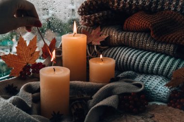 Merhaba güz, kış tatilini pencerenin kenarındaki evde kutluyoruz Şükran Günü atmosferinde Şükran Günü yaprakları, baharatlar ve sıcak sarı ışıkta örülmüş sıcak süveter üzerine mum. Pencerenin dışında Seçici Odaklanma Yağmuru