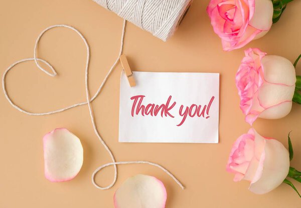 Нежные розовые розы с катушкой белой хлопковой веревки в форме сердца на бежевом фоне. Бумажная записка с текстом спасибо. Нейтральные тоны земли. Поздравительные открытки. Романтические пастельные розовые цветы