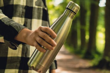 Tanımlanamayan bir kadının su şişesini tuttuğu yakın plan. Parkta yeniden kullanılabilir çelik termo su şişesi. Sürdürülebilir yaşam tarzı. Plastiksiz sıfır atıksız yaşam. Yeşil Çevre Koruma 'ya git