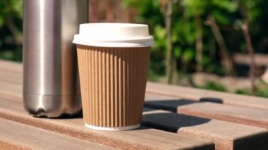 Tahta bankta kağıt bardakla yeniden kullanılabilir çelik termo su şişesi. Sürdürülebilir yaşam tarzı. Plastiksiz sıfır atıksız yaşam. Yeşil çevre korumasına geçin. Sağlık bilinci yerinde. Çelik termo su