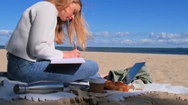 Deniz manzarasının yanındaki plajda not defteriyle ders çalışan genç bir öğrenci. Kahve içip kruvasan yemek. Minnettarlık günlüğü yazarken, kendini keşfetme, açık havada, sonbahar sıcağında.. 