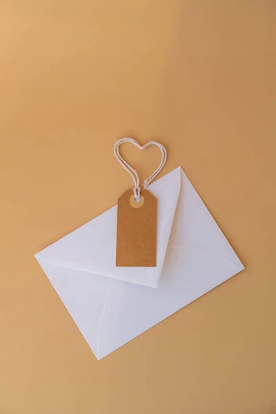 White envelope with beige paper note and rope in heart shape on neutral beige background. Greeting card holiday idea. Mock up copy space for text. Gift present