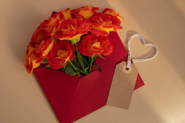 Beautiful red roses flowers in postal red envelope on red background, beige paper note and rope in heart shape copy space for text, spring time, greeting card for holiday. Flower delivery. Delicate