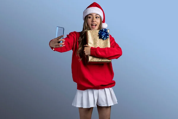 Genç Latin bir kadın yılbaşı kıyafetleriyle bir hediyeye sarılıyor telefonuyla selfie çekiyor.