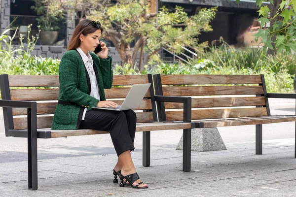 Femeie tânără vorbește la telefon în timp ce lucrează pe laptopul ei stând pe o bancă de stradă — Fotografie, imagine de stoc