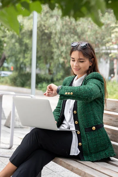 O femeie își verifică ceasul inteligent în timp ce lucrează pe laptopul ei stând pe o bancă de lemn în stradă — Fotografie, imagine de stoc