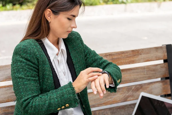 O femeie își verifică ceasul inteligent în timp ce lucrează pe laptopul ei stând pe o bancă de lemn în stradă — Fotografie, imagine de stoc