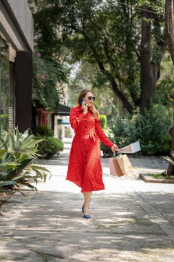 Kırmızı elbiseli genç bir kadın cep telefonuyla konuşurken alışveriş çantalarıyla sokakta yürüyor.