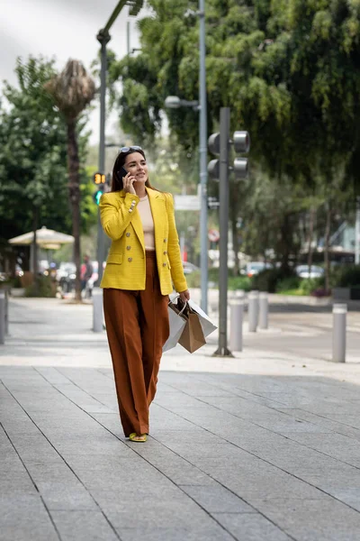 Tânăra femeie fericită mergând pe stradă cu saci de cumpărături în timp ce vorbea pe telefonul mobil — Fotografie, imagine de stoc
