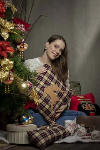 Junge kaukasische Frau lächelt neben einem Weihnachtsbaum mit einem Weihnachtskissen in der Hand — Stockfoto