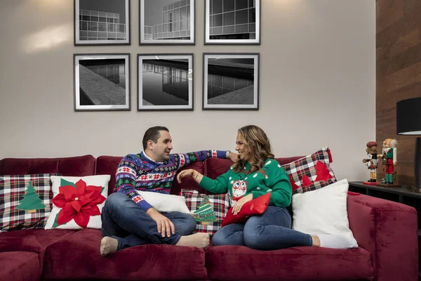 Glückliches hispanisches Paar hängt an Heiligabend auf der Couch und trägt Weihnachtspullover — Stockfoto
