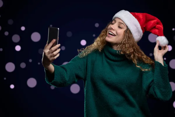 Junge lateinamerikanische Frau mit Weihnachtsmütze macht ein fröhliches Selfie mit einem Hintergrund aus unkonzentrierten Lichtern — Stockfoto