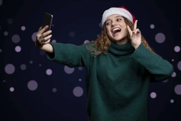 Frau in Pullover und Weihnachtsmütze macht Liebes- und Friedenszeichen, während sie ein Selfie macht — Stockfoto