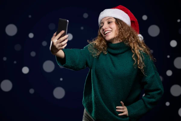 Junge Lateinerin lächelt, trägt Pullover und Weihnachtsmütze und macht ein Selfie mit ihrem Handy — Stockfoto