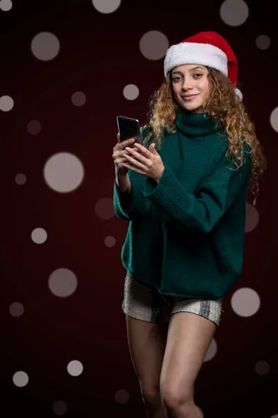 Junge Frau mit Kleidung und Weihnachtsmütze hält ein Handy in der Hand, während sie in die Kamera blickt — Stockfoto