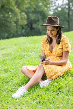 Young Mexican woman with yellow dress and sombrero sitting on the grass checking her cell phone